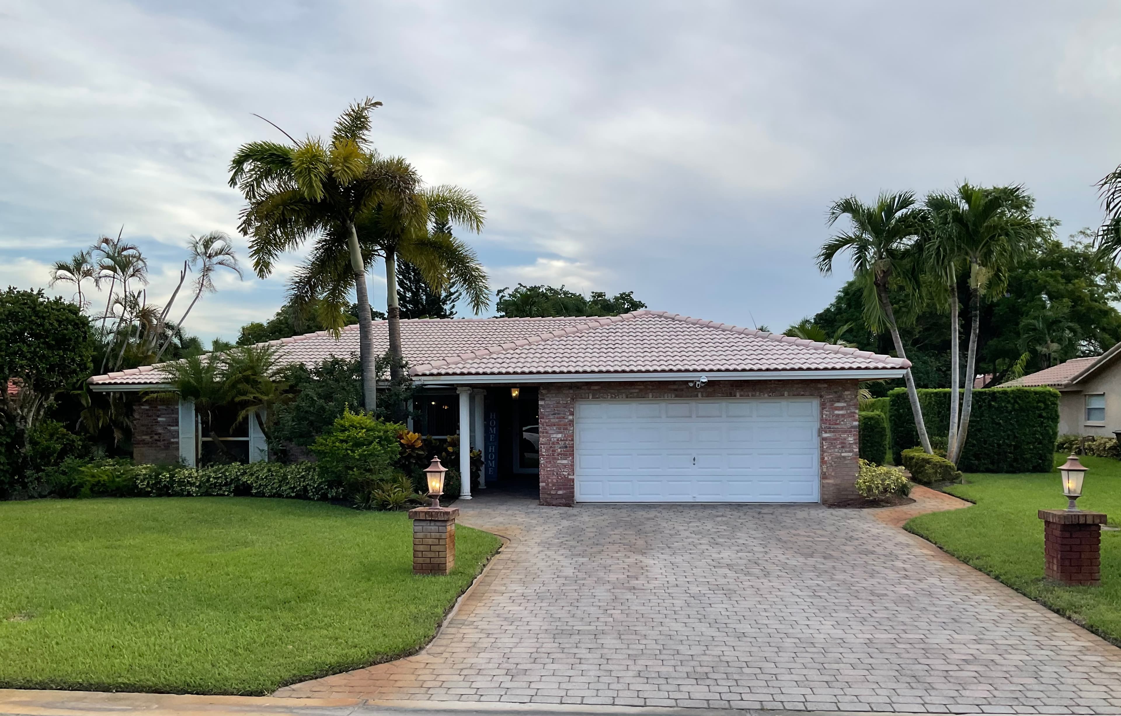 Single-story house with pink tiled roof, white garage, and paved driveway surrounded by palm trees.