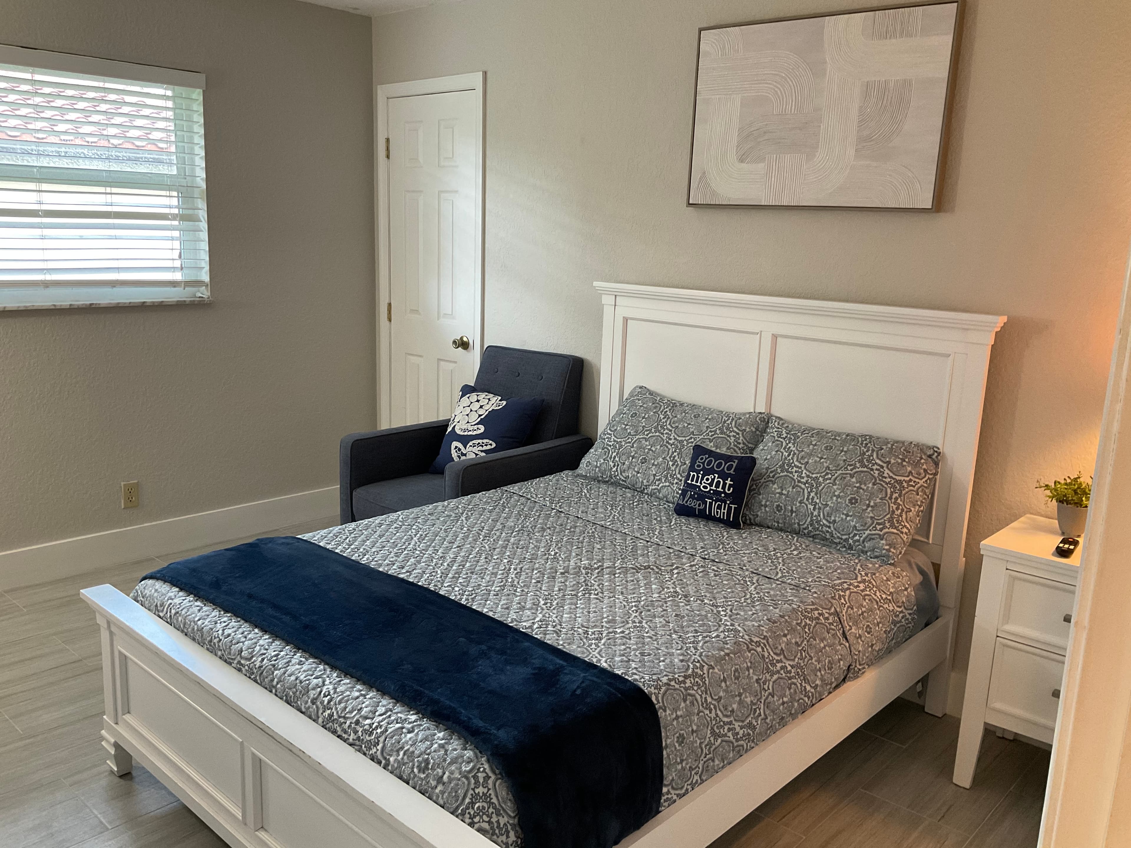 White bed with grey patterned bedding and navy accents next to a blue armchair.