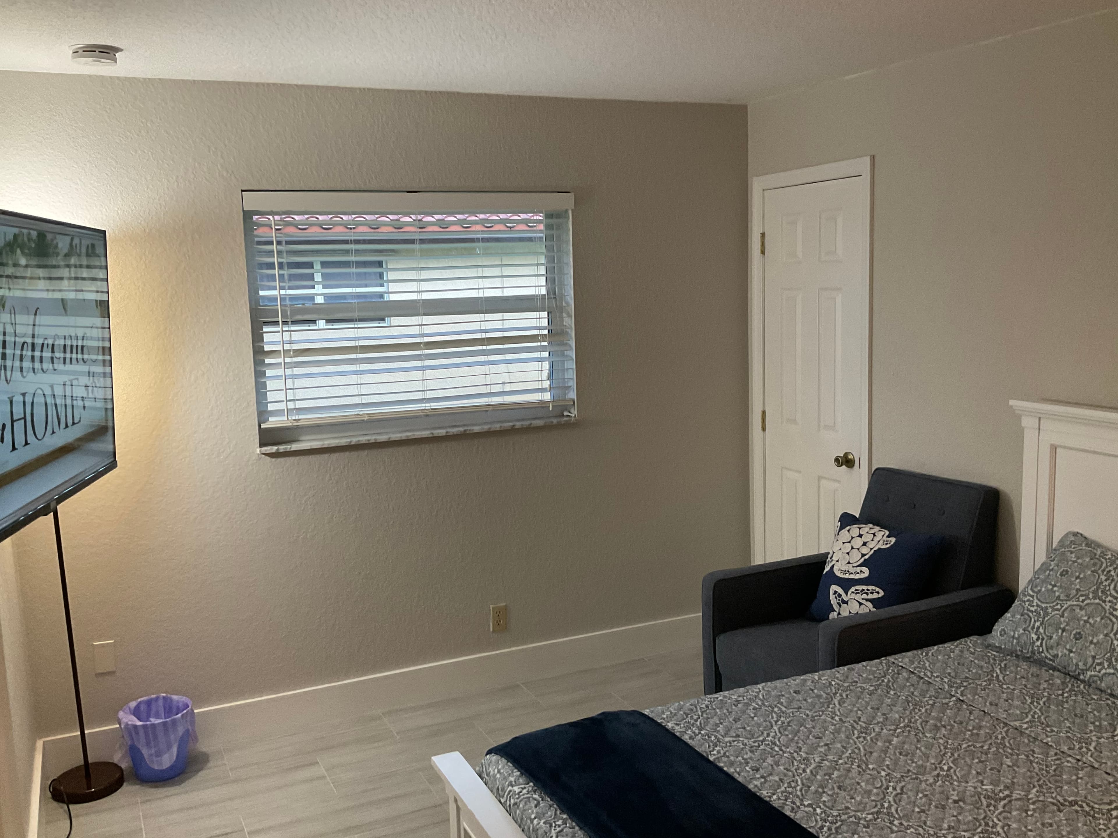 Bedroom featuring a grey patterned bed, blue armchair, and TV displaying "Welcome HOME" text.
