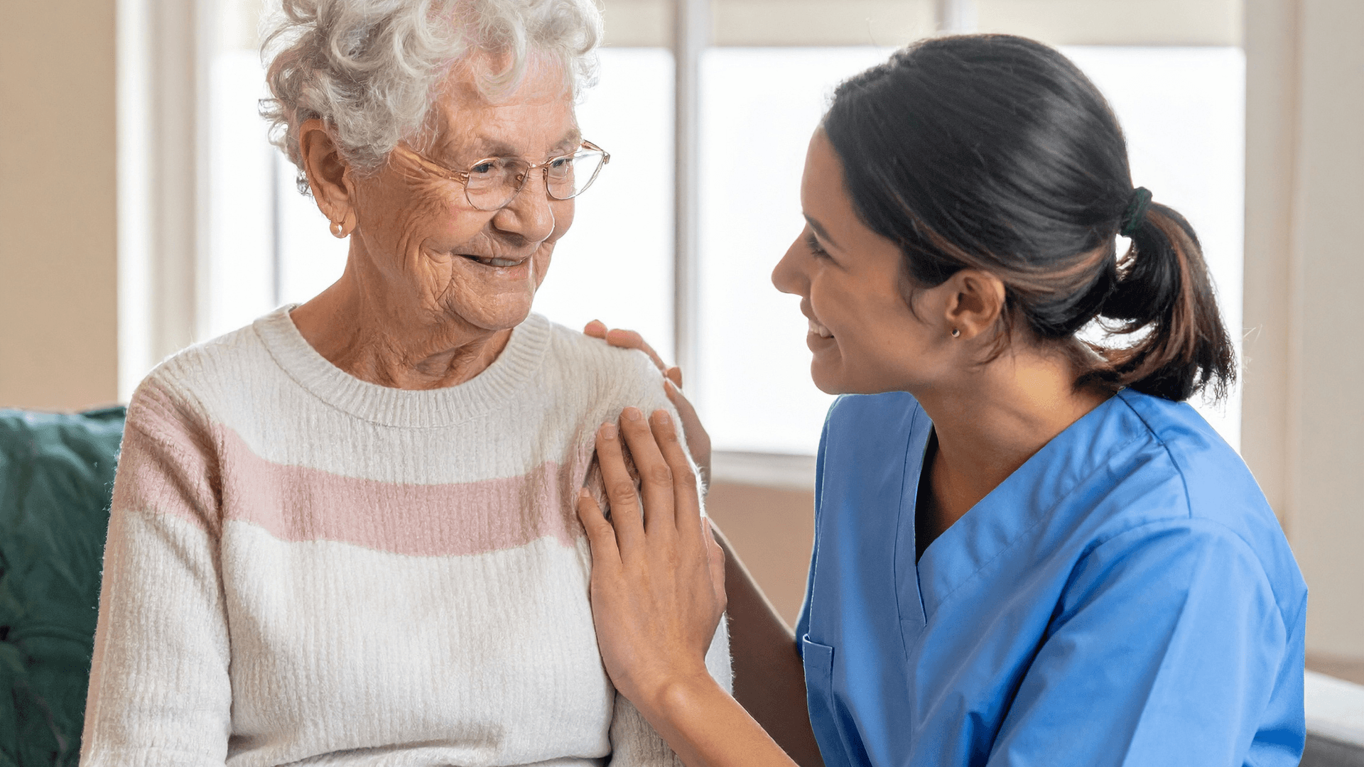 Smiling nurse in blue scrubs comforts an elderly woman with white hair and glasses.